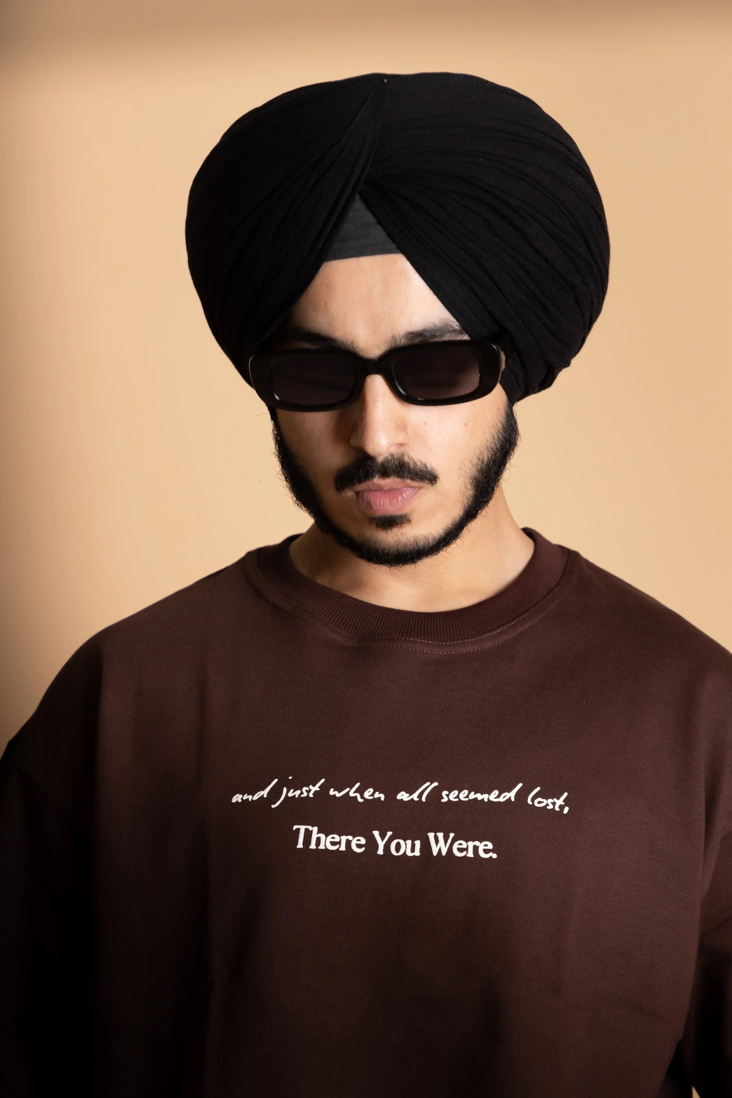 A man wearing brown oversized T-shirt showing the front puff print text.